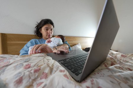 Photo for Mother lies in bed, working on her laptop with her newborn baby resting on her chest. This scene highlights the blend of professional and personal life, symbolizing the contemporary role of mothers. - Royalty Free Image