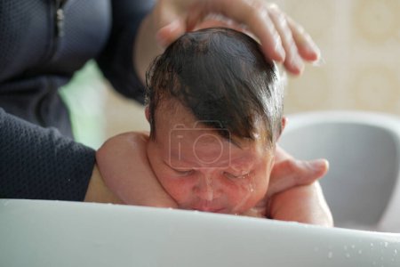 Un nouveau-né est baigné doucement dans un bassin blanc, soutenu par la main d'un parent. Le bébé semble serein, profitant de l'eau apaisante et des soins tendres pendant cette heure de bain intime et apaisante