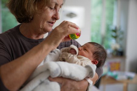 Foto de Grandmother holding a newborn baby wrapped in a white blanket and playing with a colorful toy rattle. The baby looks curiously at the rattle, creating a moment of playful interaction and bonding. - Imagen libre de derechos