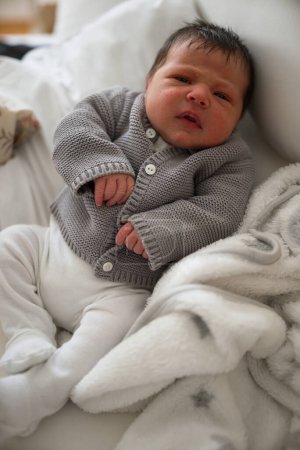 Neugeborenes Baby liegt auf einem weichen Bett in grauer Strickjacke und weißer Hose, mit offenen Augen und wachem Gesichtsausdruck und fängt einen Moment früher Neugier in einer gemütlichen häuslichen Umgebung ein
