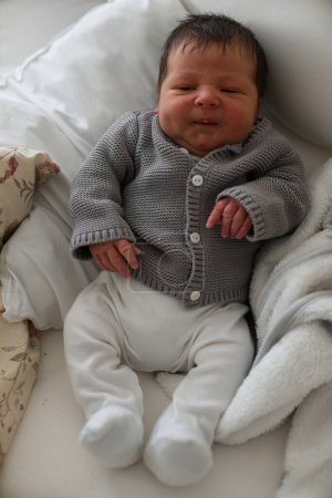 Photo for Newborn baby in a gray knitted cardigan and white pants, lying on a soft bed, showing a slight smile, capturing a peaceful moment of early life in a warm and comfortable home environment - Royalty Free Image