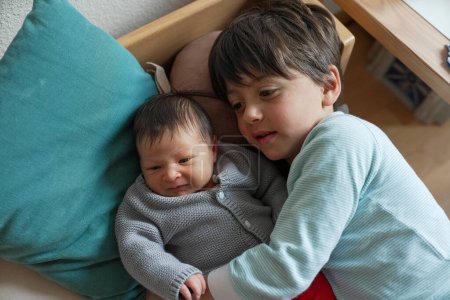 Older sibling cuddling with newborn baby, both nestled on soft pillows. The cozy and intimate setting, with natural light, highlights the strong sibling bond and love
