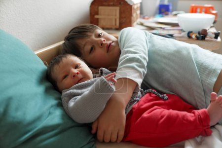 Foto de Hermano mayor abrazando al bebé recién nacido en la cama, compartiendo un momento dulce y tierno. La suave iluminación y la acogedora ropa de cama se suman al cálido y cariñoso ambiente familiar - Imagen libre de derechos