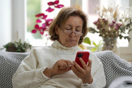 Mujer anciana en gafas con un suéter blanco sentada en un sofá, absorta en su teléfono inteligente, en una habitación acogedora y bien iluminada