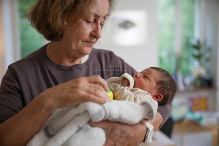 Foto de Grandmother gently holding her newborn grandchild wrapped in a white blanket, engaging the baby with a colorful toy rattle. The baby is focused on the rattle, capturing a moment of playfulness - Imagen libre de derechos