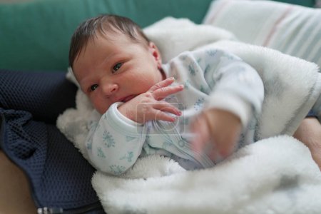 Ein Neugeborenes kuschelte sich in eine weiße Decke und trug einen weichen Einteiler. Das Baby ist ruhig und aufmerksam und beobachtet die Umgebung mit einem friedlichen und neugierigen Gesichtsausdruck