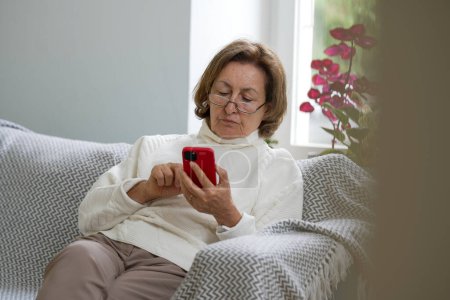Photo for A senior woman wearing glasses and a white sweater is seated on a couch, deeply engaged with her smartphone, emphasizing a warm and cozy moment in a well-lit room - Royalty Free Image