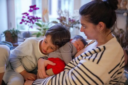 Mother cradling her newborn baby with an older child lovingly snuggling up to both of them. The room is softly lit with a warm, intimate ambiance that emphasizes family bond