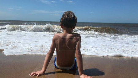 Young boy at the beach, seated waiting for ocean waves