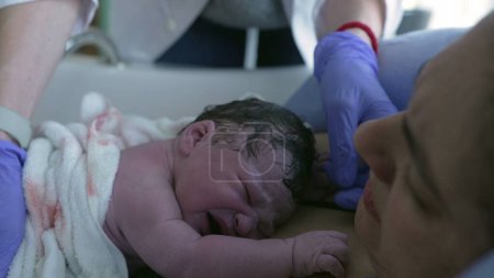 Doctor inspecting newborn baby during initial seconds of life crying on mother's chest, feeling mom's skin right after giving birth, first cries of infant during childbirth