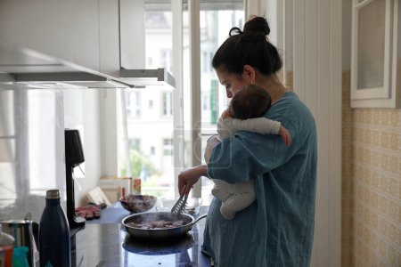 Téléchargez les photos : Femme en chemise bleue tenant bébé dans la cuisine, multitâche entre le rôle parental et les tâches quotidiennes, illustrant l'équilibre entre la maternité et les responsabilités ménagères dans une maison moderne - en image libre de droit