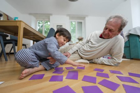 Älterer Mann und kleiner Junge spielen ein Memory-Spiel, verbinden sich über eine einnehmende Aktivität, Innenraumszene mit natürlichem Licht, Hervorhebung der familiären Verbundenheit und der Interaktion zwischen den Generationen, lustiger Moment