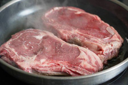 Ribeye steaks in a pan starting to sear, emphasizing the marbling and freshness of the meat,