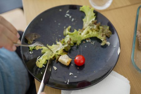Nahaufnahme eines schwarzen Tellers mit Salatresten und einer Kirschtomate, die das Ende einer Mahlzeit darstellt und einfache, gesunde Essgewohnheiten und das tägliche Essen in einem ungezwungenen häuslichen Umfeld repräsentiert