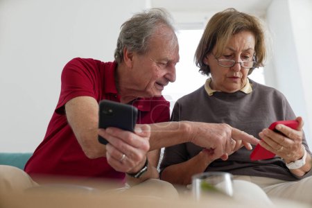 Elderly couple using smartphones, man pointing at the woman's screen, indoor setting with bright natural light, representing modern communication and technology use among senior citizens