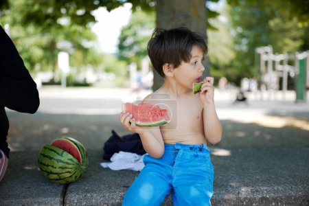 Ein kleiner Junge in blauer Hose sitzt auf einer Parkbank und genießt an einem sonnigen Tag ein Stück Wassermelone. Die saftigen Früchte, die Konzentration des Kindes und die sommerliche Kulisse schaffen einen perfekten Moment kindlicher Freude