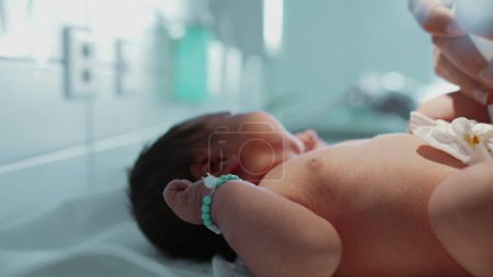 Säugling mit individuellem Armband, das auf dem Krankenhausbett ruht, Daumen lutscht, zufrieden schaut, Gesicht und Hand in Nahaufnahme, Neugeborenes fängt herzerwärmende erste Momente ein