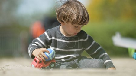Young Boy Playing with Colorful Toy Trucks in Sand at Park - Outdoor Playtime in Sweater