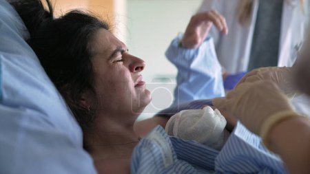 Mutter zittert vor Schmerzen direkt nach der Geburt ihres Neugeborenen, Arzt und Krankenschwestern umgeben sie mit Pflege in der Geburtsklinik, Hände legen Säugling sanft in die Brust