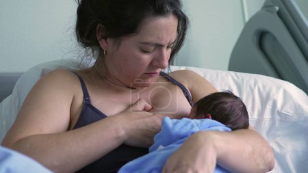 Mother breastfeeding newborn baby in a blue onesie, sitting in a hospital bed, showcasing a nurturing and intimate moment between mother and child in a postnatal care setting