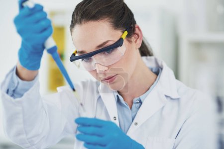 Auf geht 's. eine fokussierte junge Wissenschaftlerin mit Schutzbrille, während sie eine Testprobe in ein schnödes Laborinnere gießt