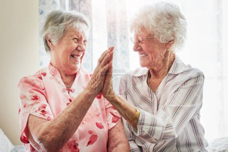 Foto de Seguimos siendo amigos después de todos estos años. dos felices ancianas dándose unos a otros una alta cinco juntos en casa - Imagen libre de derechos