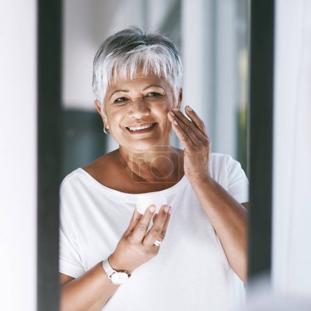 Photo for My skin looks so radiant with this on. a cheerful mature woman applying skin cream on her face while looking into a mirror at home - Royalty Free Image