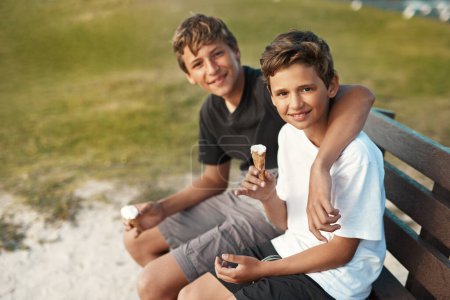 Téléchargez les photos : Tu ne peux pas acheter le bonheur, mais tu peux acheter de la glace. Portrait de deux frères heureux mangeant des cônes de crème glacée assis sur un banc près de la plage - en image libre de droit