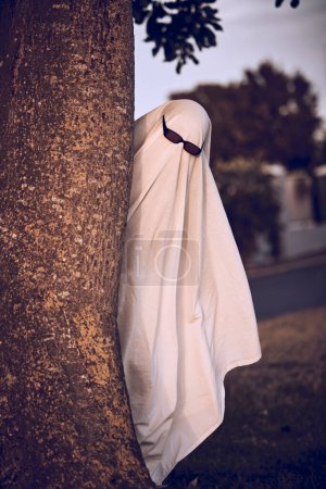 ghost costume, halloween and sunglasses in nature park outdoors. Funny horror dress up, spooky holiday celebration and creative scary mystery spirit design or hiding ghoul with glasses for carnival.