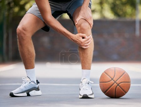 Pain, basketball and man with knee injury standing on outdoor court, holding leg. Sports, fitness and athlete with joint pain, injured and hurt in training, workout and game on basketball court.