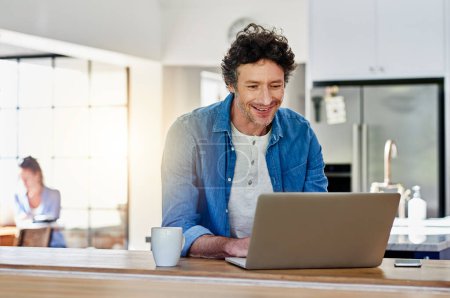 Surfs up. a happy man using his laptop while sitting in the kitchen