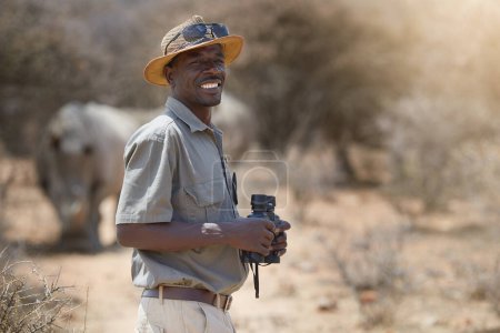 Téléchargez les photos : Je vis mon rêve de carrière. Portrait d'un garde forestier confiant regardant un groupe de rhinocéros dans le veld - en image libre de droit