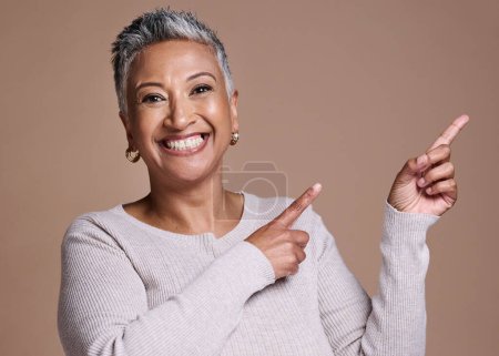 Photo for Woman, smile and happiness in studio for advertising free space, promo or mockup on a brown background for cosmetics, makeup or fashion. Face portrait of a mature model pointing finger at mock up. - Royalty Free Image
