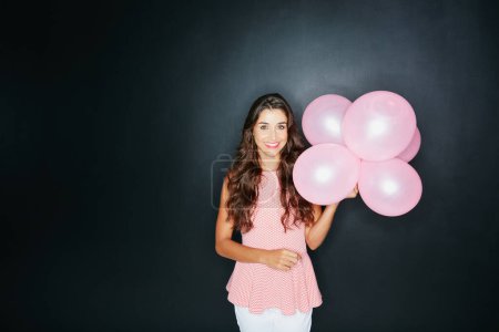 Ill bring the party to you. Studio portrait of an attractive young woman holding balloons against a dark background