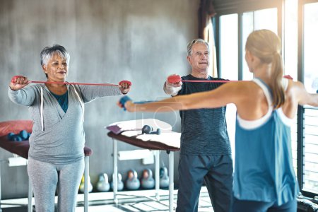 Living fit helps keep their physical functions in check. a senior man and woman using resistance bands with the help of a physical therapist