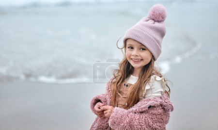 Téléchargez les photos : Fille heureuse enfant sur la plage, portrait pendant les vacances d'hiver avec bonnet rose et sourire enfant sur le bord de mer de Dublin. Liberté en plein air sur la pause océan, tout-petit mignon relaxant au bord de l'eau et la paix côtière. - en image libre de droit