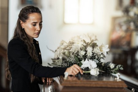 Beerdigung, traurig und Frau mit Blumen auf dem Sarg nach dem Verlust eines geliebten Menschen, Familie oder Freundes. Trauer, Tod und junge Frau legen eine Rose auf Sarg in der Kirche mit Traurigkeit, Depression und Trauer.