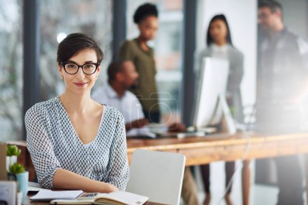 Photo for Plans have been progressing well so far. Portrait of a young businesswoman sitting at her desk with colleagues in the background - Royalty Free Image