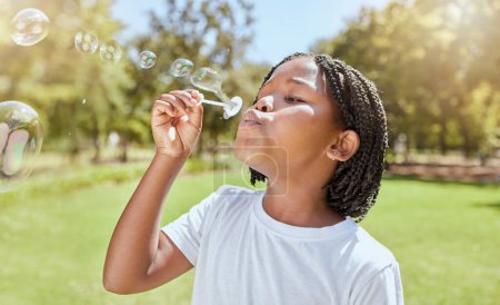 Téléchargez les photos : Parc, enfant et fille noire soufflant des bulles profiter de plaisir seul à l'extérieur, la joie et le développement de l'enfance. Joyeux, liberté et enfant apprendre et jouer avec un jouet à bulles de savon ou baguette et se détendre sur l'herbe. - en image libre de droit