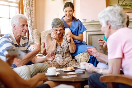Téléchargez les photos : Au vainqueur va le butin. un groupe d'aînés heureux jouant à un jeu de cartes dans leur maison de retraite pendant qu'une infirmière regarde - en image libre de droit