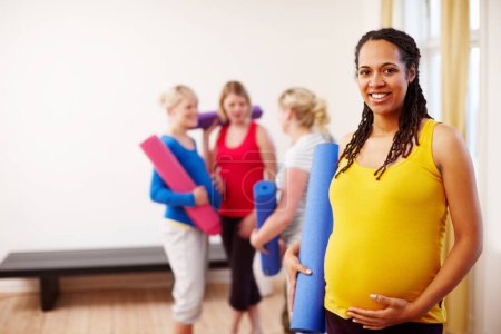 Photo for Relaxation exercise is perfect for pregnancy. A happy young pregnant African-American woman holding her exercise mat while standing in a gym with her friends in the background - Royalty Free Image