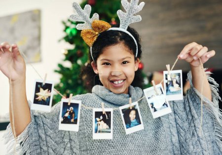 Téléchargez les photos : Noël, célébration et fille avec polaroid pour la décoration, heureux et montrer des photos dans la maison. Sourire, excité et portrait d'un enfant avec des photos pour décorer la maison pour un Noël festif. - en image libre de droit
