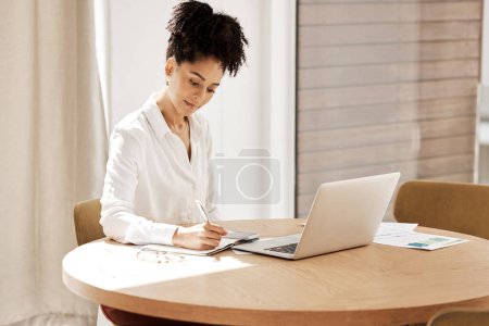 Woman writing in notebook, laptop on table and home office of accountant, auditor or financial advisor. Finance report, strategy and budget planning, black woman doing research on tax audit documents.