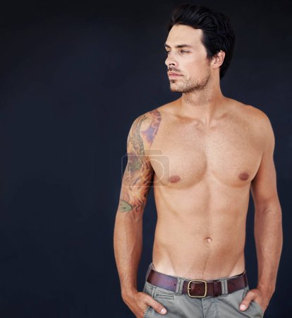 Photo for His tattoo brings an edge to his boyish good looks. handsome male with his shirt off with a dark background - Royalty Free Image