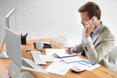 Hes a capable investment manager. a young businessman talking on the phone while writing at his desk