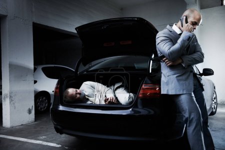 Foto de Indefenso frente a un crimen violento. Un hombre de negocios asustado yaciendo en el maletero de un coche mientras su captor habla por un teléfono celular - Imagen libre de derechos