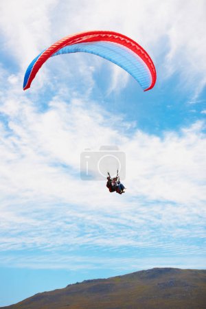 Photo for Over the hills...Low angle view of two people doing tandem paragliding high up with a hill in the background - Royalty Free Image