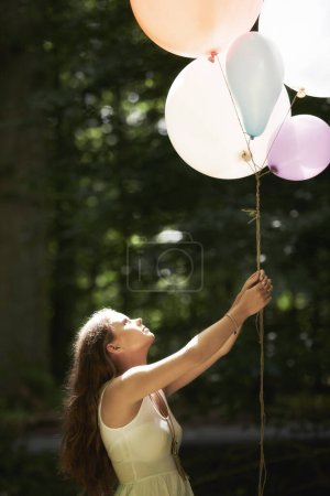 A pretty young woman holding a bunch of balloons in the forest.