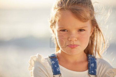 Blonde caucasian little girl with pouted lips looking angry, annoyed and stubborn while spending a summer day at the beach. Cute kid expressing frustration and throwing a tantrum. Naughty bored child.