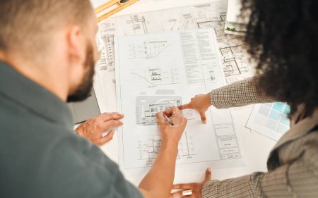 Architects working on a blueprint from above. Businesspeople writing on a building plan. Coworkers working on architect plan. Businessman working with a businesswoman in his office